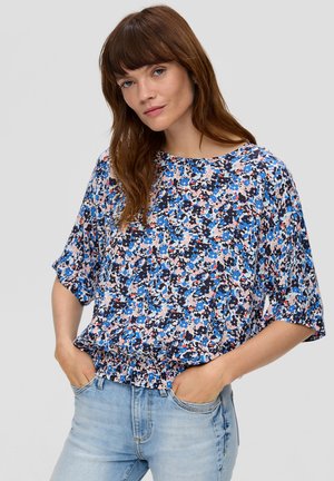 Blumenbluse mit einer Mischung aus blauen, pinken und weißen Mustern, lockerer Passform mit kurzen Ärmeln und gerafftem Saum. Kombiniert mit hellen Jeans.