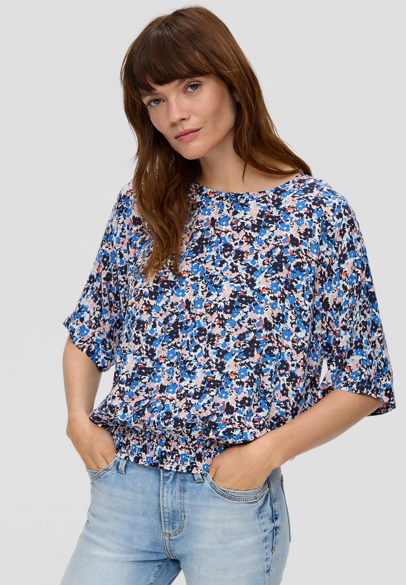 Bloemige blouse met een mix van blauwe, roze en witte patronen, een losse pasvorm met korte mouwen en een geplooide onderkant. Gecombineerd met lichte spijkerbroeken.