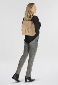 Sac à dos en cuir beige avec fermeture à cordon et rabat, associé à un jean gris et des talons noirs, présenté de côté sur un fond clair.