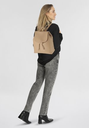 Sac à dos en cuir beige avec fermeture à cordon et rabat, associé à un jean gris et des talons noirs, présenté de côté sur un fond clair.