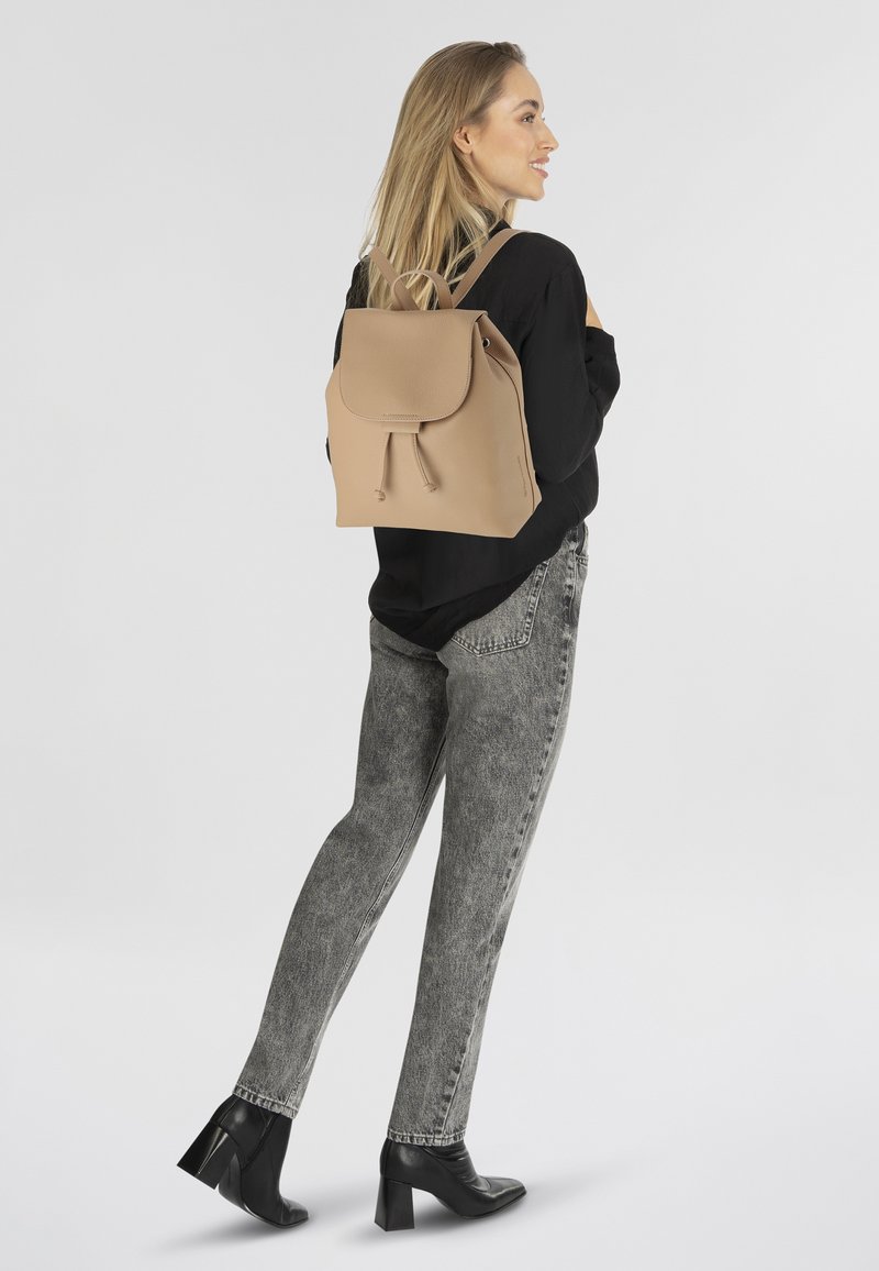 Beige leren rugzak met trekkoordsluiting en klep, gecombineerd met grijze jeans en zwarte hakken, zijaanzicht tegen een lichte achtergrond.