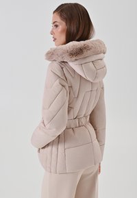 Vrouw in een gebreide beige winterjack met een muts met bontafwerking, gezien vanuit de zij- en achterkant tegen een effen achtergrond.