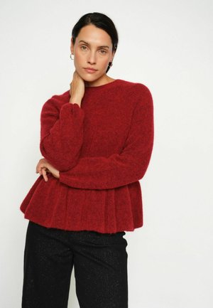 Roter Strickpullover mit texturiertem Finish, rundem Ausschnitt und peplum Saum. Kombiniert mit schwarzen glitzernden Hosen.