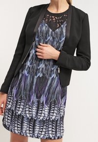 Blazer noir porté sur une robe sans manches avec un motif de plumes en violet, bleu et noir, avec un détail de décolleté en dentelle.