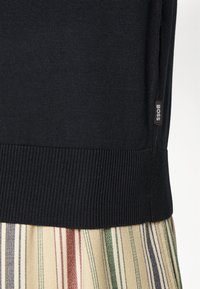 Černý svetr s ribbovaným spodním lemem, částečně viditelný přes pruhovanou sukni s béžovými, zelenými a červenými pruhy. Viditelná značka s logem BOSS.