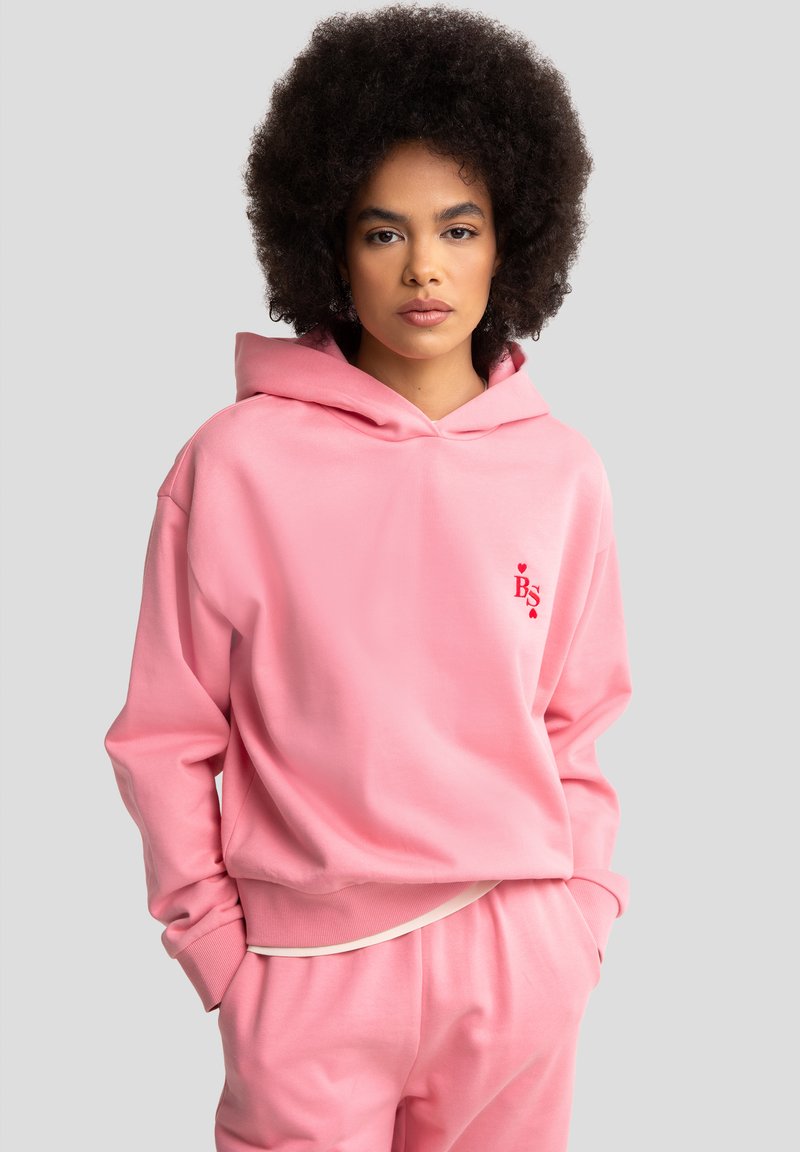 Hoodie rose en tissu doux avec une poche kangourou. Comprend une capuche, des poignets côtelés et un petit logo rouge sur la poitrine.
