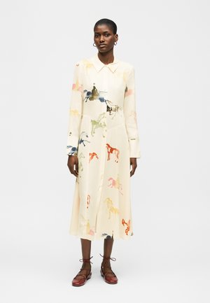 Γυναίκα που φοράει μπεζ midi φόρεμα με μακριά μανίκια και πολύχρωμα σχέδια αλόγων, καθώς και μπορντό δερμάτινα παπούτσια με κορδόνια, στέκεται μπροστά σε έναν απλό φόντο.