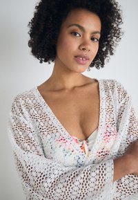 Cubierta de ganchillo blanca con un escote en V profundo, con mangas sueltas. Un patrón floral asoma por debajo, añadiendo color y detalle.
