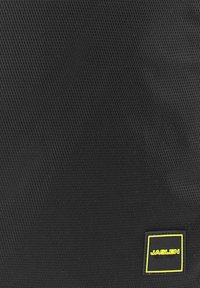 Bolsa textil negra con un patrón de malla, que presenta un logo cuadrado en amarillo y negro. El material tiene un acabado texturizado.