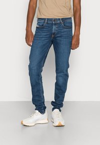 Homem vestindo jeans justo azul e ténis bejes, de pé contra um fundo claro e simples.