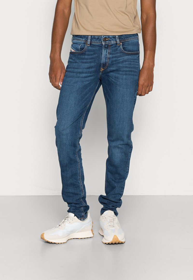 Homem vestindo jeans justo azul e ténis bejes, de pé contra um fundo claro e simples.