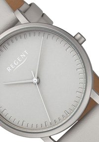Montre en métal argenté avec un cadran rond minimaliste, un cadran gris clair et des aiguilles des heures et des minutes fines. Bracelet en cuir marron avec des accents argentés.