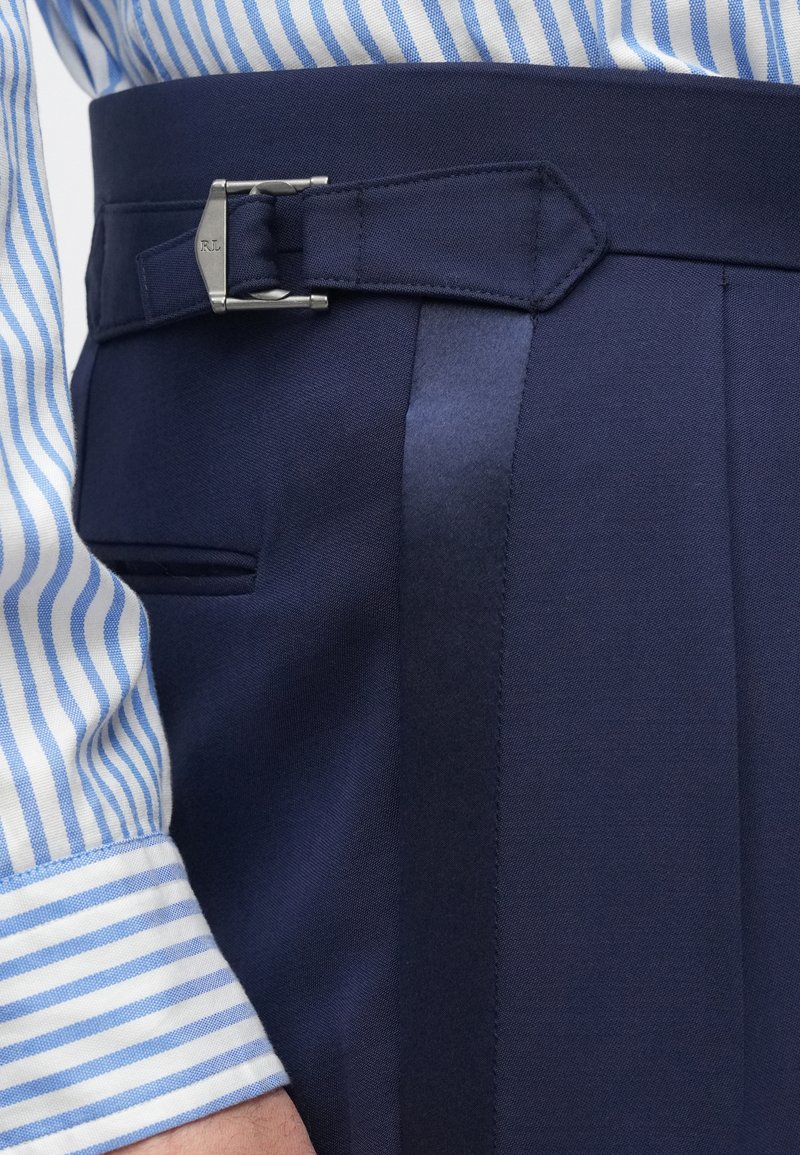 Navy trousers with a satin stripe, featuring a silver metal buckle and pleated design. Blue and white striped shirt cuff visible.
