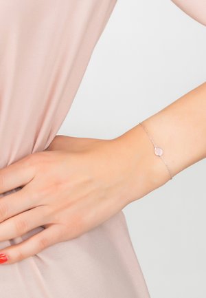 Bracciale a catena d'argento con un gemstone rosa a forma di goccia, indossato su una mano contro uno sfondo di morbido tessuto rosa.
