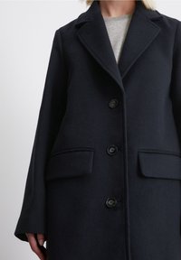 Abrigo de lana azul marino con cuello con muescas, tres botones y dos bolsillos delanteros. La tela tiene una textura suave y un diseño limpio y estructurado.