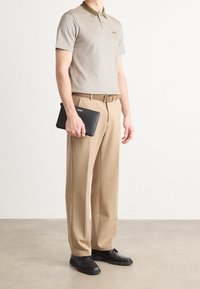 Polo beige avec un col vert et un logo, pantalon beige, ceinture noire, tenant une tablette noire et portant des chaussures noires.