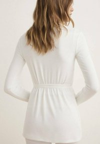 Top blanc à manches longues avec une taille cintrée et une texture côtelée, présentant un dos lisse et un design ceinturé subtil.