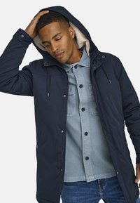 Marinblå parka med huva, med en mjuk foder, två framfickor och knäppningar med knappar. Bärs över en ljusblå denimskjorta.