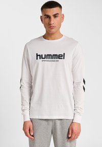 Biała koszulka z długim rękawem z czarnym logo "hummel" i napisem "SPORTSWEAR SINCE 1923". Posiada czarne paski na rękawach. Gładki bawełniany materiał.