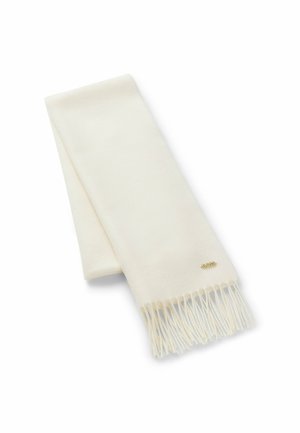 Zachte crèmekleurige sjaal met franjes aan de uiteinden en een klein goudkleurig merkje dicht bij één rand, opgevouwen liggend op een witte achtergrond.