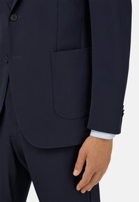 Marynarka w kolorze granatowym z jedną kieszenią na piersi i dwoma guzikami, o gładkiej fakturze, z kontrastową, jasnoniebieską koszulą w paski pod spodem.