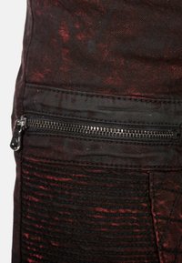 Denim nero con finitura consumata, caratterizzato da dettagli rossi. Include una tasca con zip e texture a coste lungo la cucitura laterale.