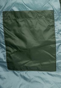 Světle modrá vnější tkanina s hladkým povrchem, vybavená obdélníkovou tmavozelenou kapsou, prošitými okraji a minimalistickým designem.