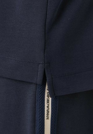 Primo piano di tessuto blu navy con dettaglio di spacco laterale ed etichetta visibile con la scritta "Emporio Armani" su striscia in rete sottostante.