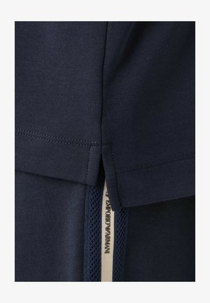 Primo piano di tessuto blu navy con dettaglio di spacco laterale ed etichetta visibile con la scritta "Emporio Armani" su striscia in rete sottostante.