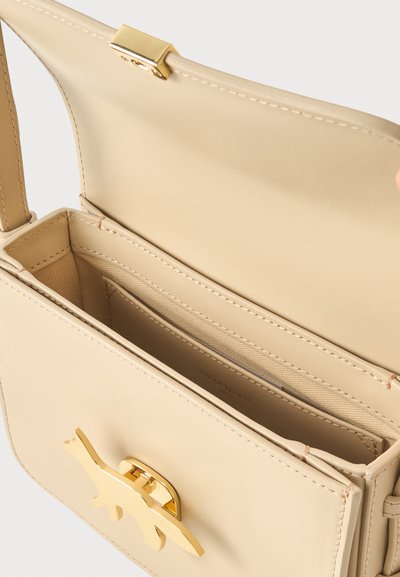 Μπεζ ορθογώνια τσάντα χειρός ανοιχτή για να δείξει τα εσωτερικά διαμερίσματα, με κούμπωμα σε σχήμα δεινοσαύρου από χρυσό και ορατό ιμάντα ώμου.