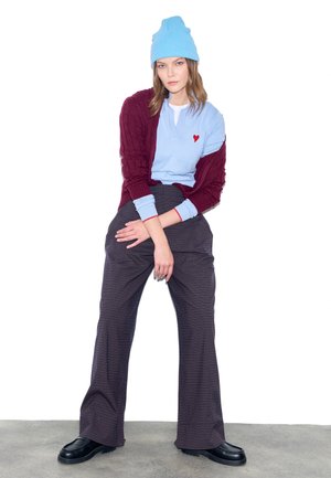 Junge Frau mit blauer Mütze, hellblauem Oberteil mit rotem Herz, burgunderfarbenem Pullover, dunkler weiter Hose und schwarzen Schuhen, vor weißem Hintergrund sitzend.