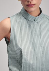 Blusa sin mangas de color azul claro con rayas verticales, cuello de pie y detalle de botones. Tejido suave con un ligero brillo.