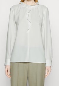 Blouse blanche à manches longues avec col à volants, texture lisse, patte de boutonnage à l'avant, et coupe décontractée. Associée à un pantalon kaki clair.