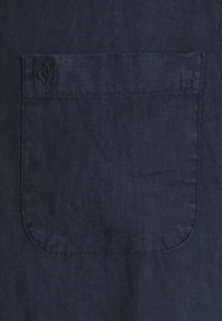 Marineblauwe linnen stof met een enkele patachzak met subtiele stiksels en een verdiept logo in de linkerbovenhoek.