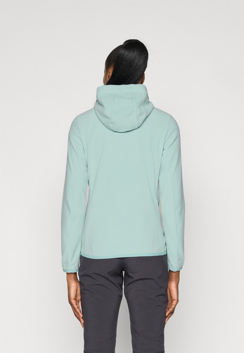 Helle türkisfarbene Fleece-Jacke mit einer gesteppten Kapuze, langen Ärmeln und einer schlanken Passform. Getragen mit dunkelgrauen Hosen. Glatte Textur.