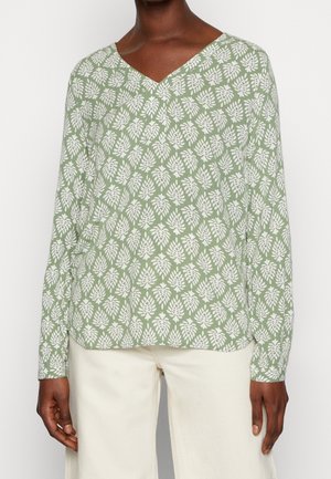 Mujer con camiseta de manga larga verde con patrón de hojas blancas y escote en V, combinada con pantalones color crema, de pie frente a un fondo blanco.