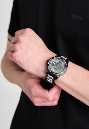 Black watch with a textured dial and silver accents, featuring a black strap with a pattern of small square links. Worn on a wrist.