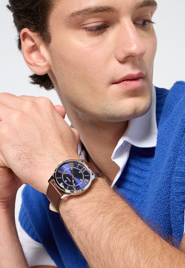 Braune Lederarmbanduhr mit blauem Zifferblatt, silbernen Akzenten und römischen Ziffern. Verfügt über ein rundes Metallgehäuse und gut lesbare Markierungen.