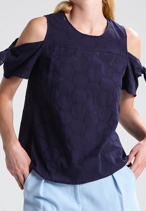 Marineblå kold skulder-bluse med cirkulært hulmønster og bundne korte ærmer, båret med lyseblå bukser.