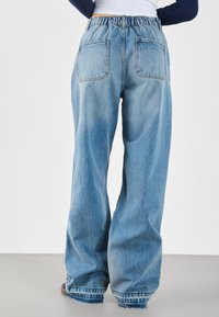Rückansicht einer Person, die eine hellblaue, weite Jeans mit elastischem Bund und sichtbaren Gesäßtaschen vor einem schlichten Hintergrund trägt.