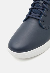 Sapatilha de couro azul-marinho com textura suave, bico arredondado, sola de borracha branca e atacadores azul-escuro sobre uma língua de malha.