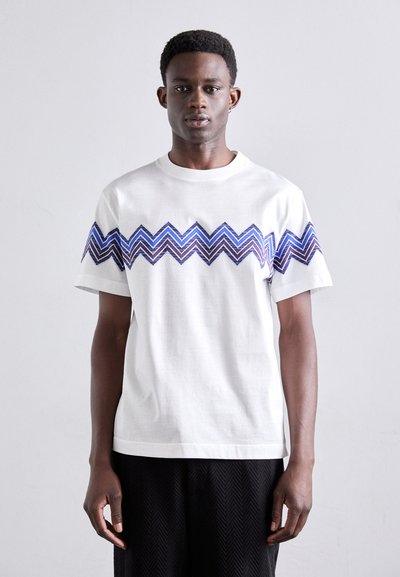 T-shirt en coton blanc avec un motif en chevrons bleu et violet sur la poitrine. Manches courtes et col rond.