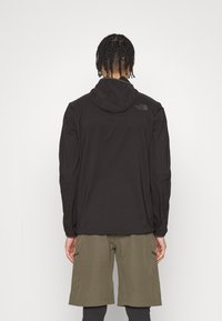 Sudadera negra ligera con capucha, que presenta un pequeño logo en la parte trasera. Combinada con pantalones cortos cargo verde oliva y leggings negros.