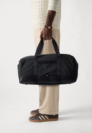 Sac de voyage noir en tissu résistant, avec des poignées robustes, une poche avant et un petit logo sur le côté. Texture lisse.