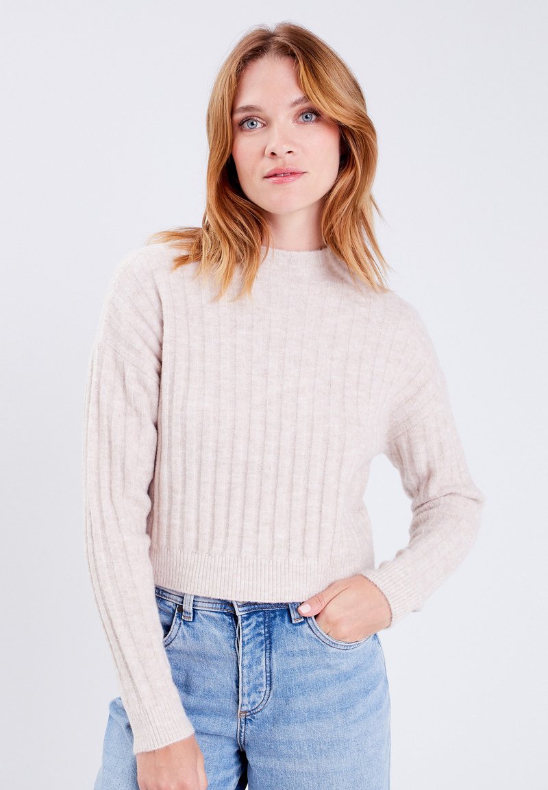 Hellbeiger, gerippter Pullover mit langen Ärmeln und hohem Ausschnitt, aus einem weichen Material gefertigt. Kombiniert mit hellblauen, hoch taillierten Jeans.