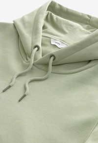 Sudadera verde menta con capucha ajustable, textura de algodón suave y una etiqueta visible en el interior. Presenta ojales de color plateado para el cordón.