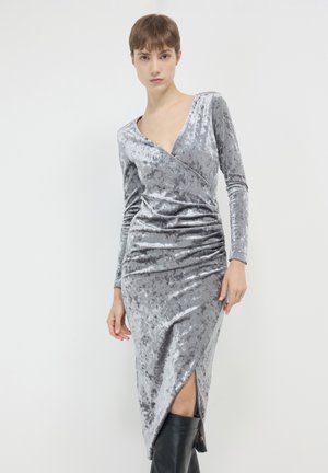 Abito in velluto grigio con scollatura a V profonda, maniche lunghe e arricciature diagonali; presenta uno spacco fino al ginocchio e una silhouette aderente.