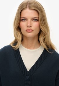 Cardigan tricoté bleu marine avec un décolleté en V, des bords côtelés et une fermeture à un bouton, porté par-dessus un haut beige clair.