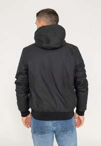 Czarna ocieplana kurtka bomberka z kapturem, ściągacze na mankietach i w pasie, gładka tekstura, o minimalistycznym designie, noszona z niebieskimi dżinsami.