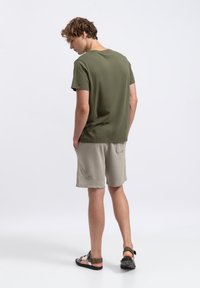 T-shirt in cotone verde oliva, vestibilità morbida, abbinata a pantaloni corti beige chiaro con tasche laterali e una superficie liscia. Sandali piatti completano il look.